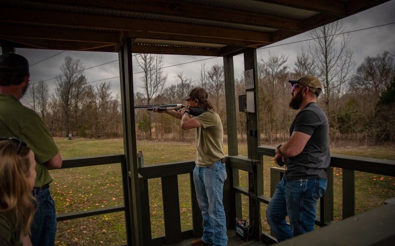 shooter standing under a shelter aiming shotgun with others around