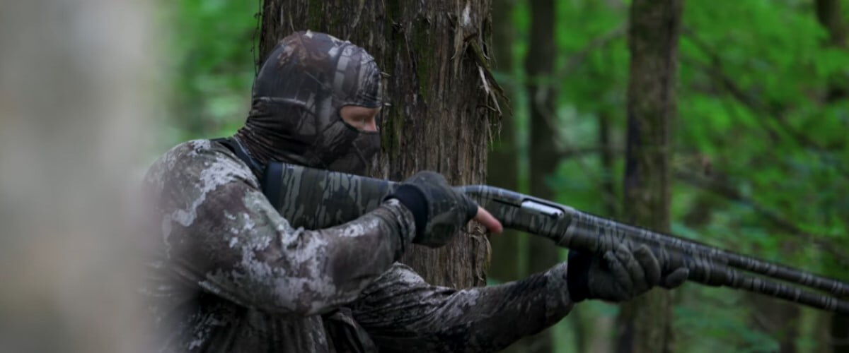 hunter wearing camo in the woods holding a shotgun