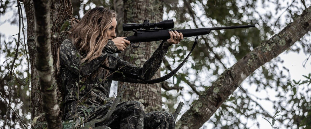Aly from Alabama sitting in a tree stand aiming a rifle