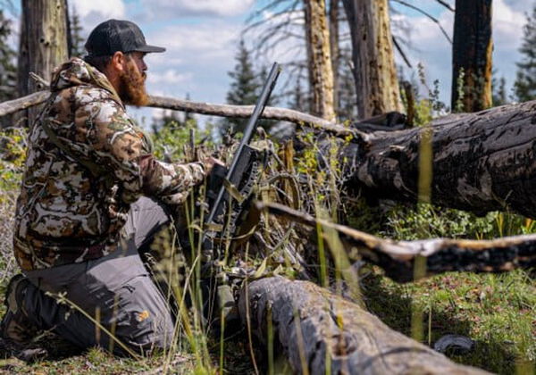 hunter kneeling behind a falling tree with his rifle leaned against it