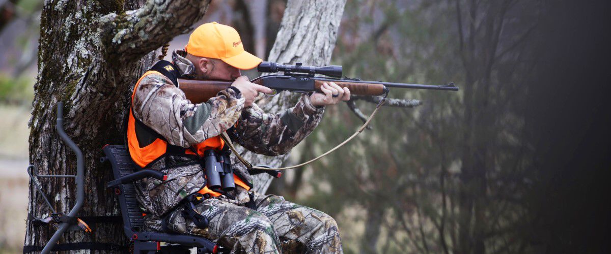 Hunter looking down the scope of a rifle while sitting in a tree stand