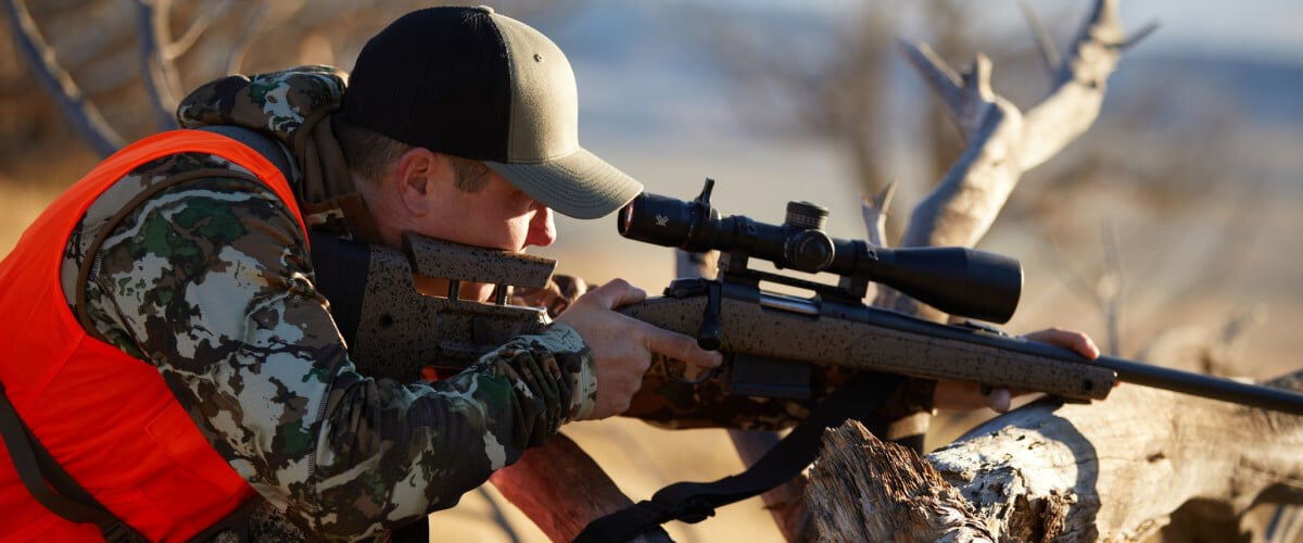 hunter looking down a rifle scope that is resting on a tree truck