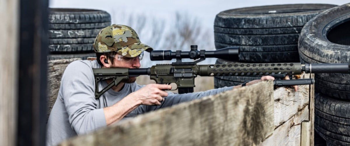 shooter looking down a rifle scope in front of a stack of tires