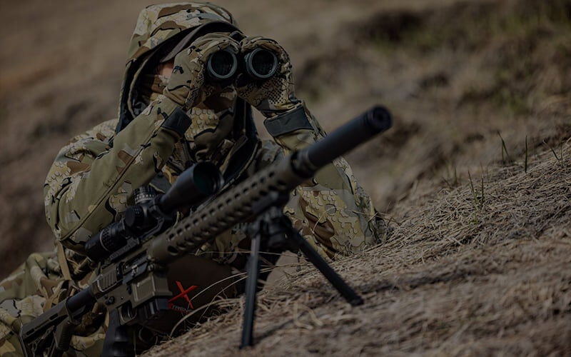 Hunter looking through binoculars while laying next to a rifle on a bipod
