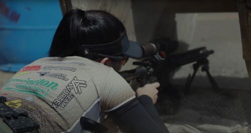 Danyelo D'Angelo laying on the group looking through the scope of a rifle pointed through an obstacle