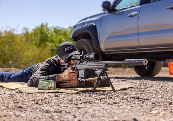 shooter laying on the ground next to a pickup and looking down the scope of a rifle