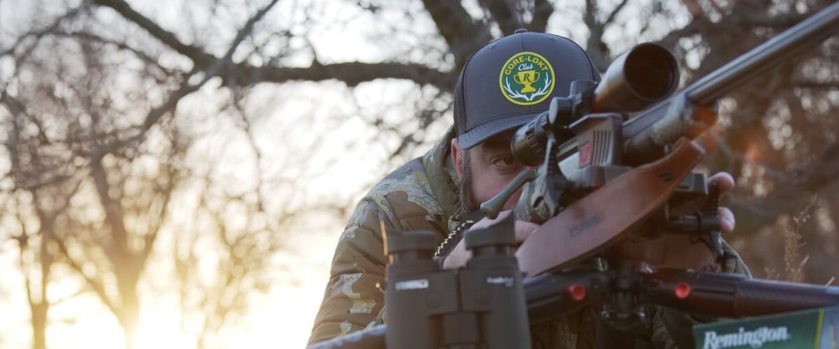 hunter looking down a rifle scope on a tri-pod with some binoculars and a box of Remington ammo