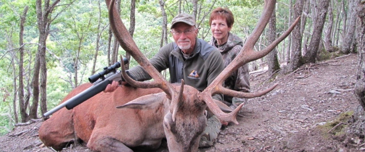 2 hunters sitting behind a dead red stag with a rifle