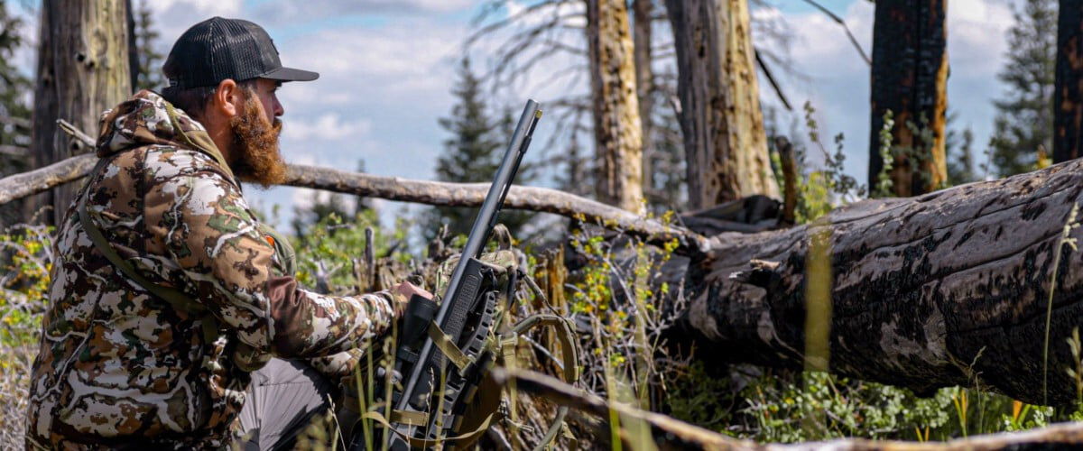 hunter kneeling behind a falling tree with his rifle leaned against it