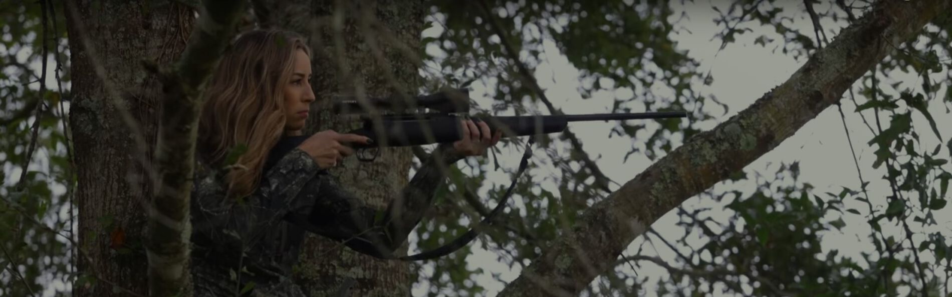 Aly looking down a rifle while in a tree stand