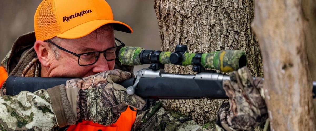 hunter looking down a rifle scope next to a tree
