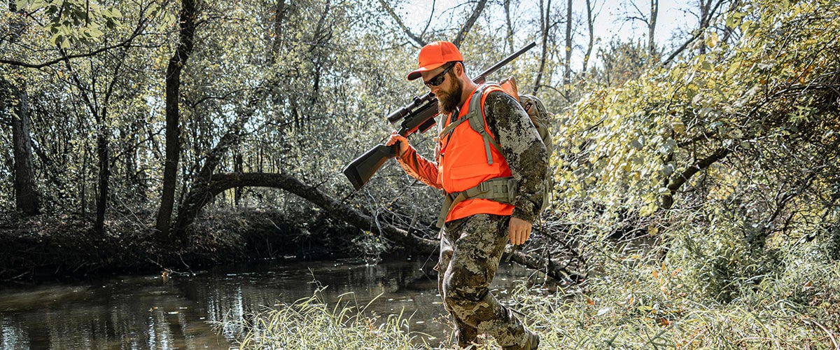 hunter walking next to a creek with a rifle on his shoulder