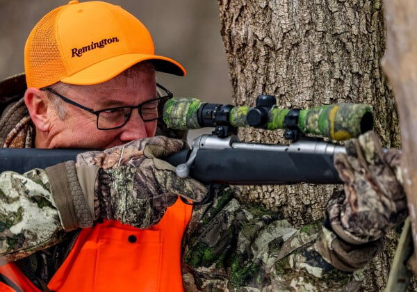 hunter looking down a rifle scope next to a tree