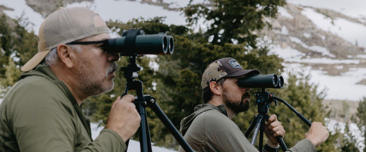 two hunters looking through monoculars while sitting on a hillside