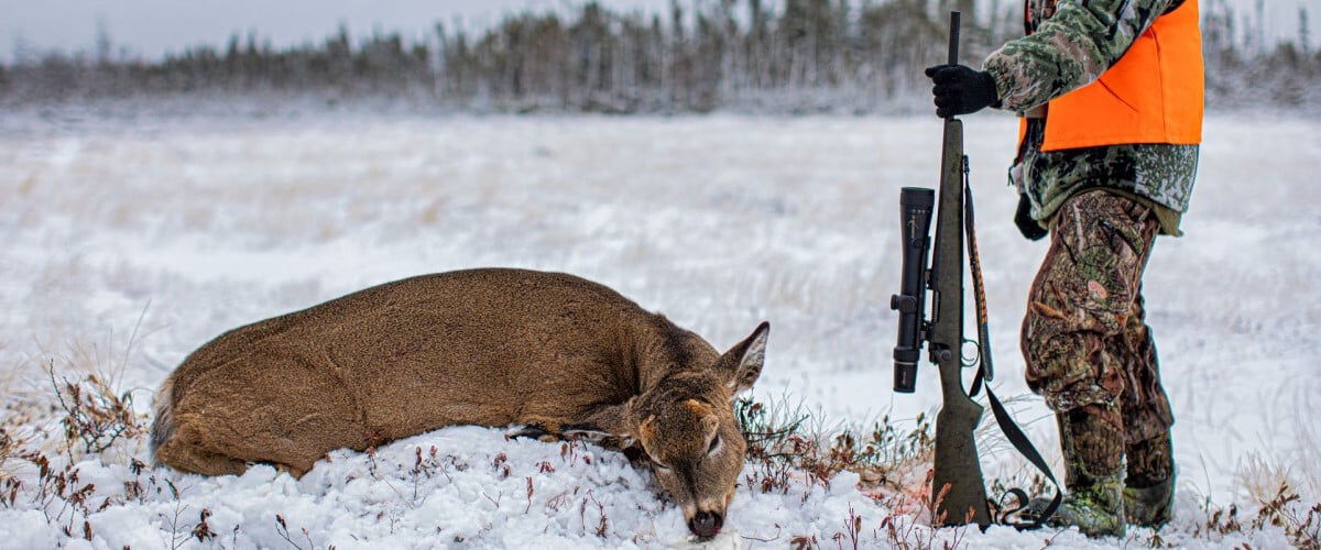 hunter standing in front of a dead deer while holding a rifle