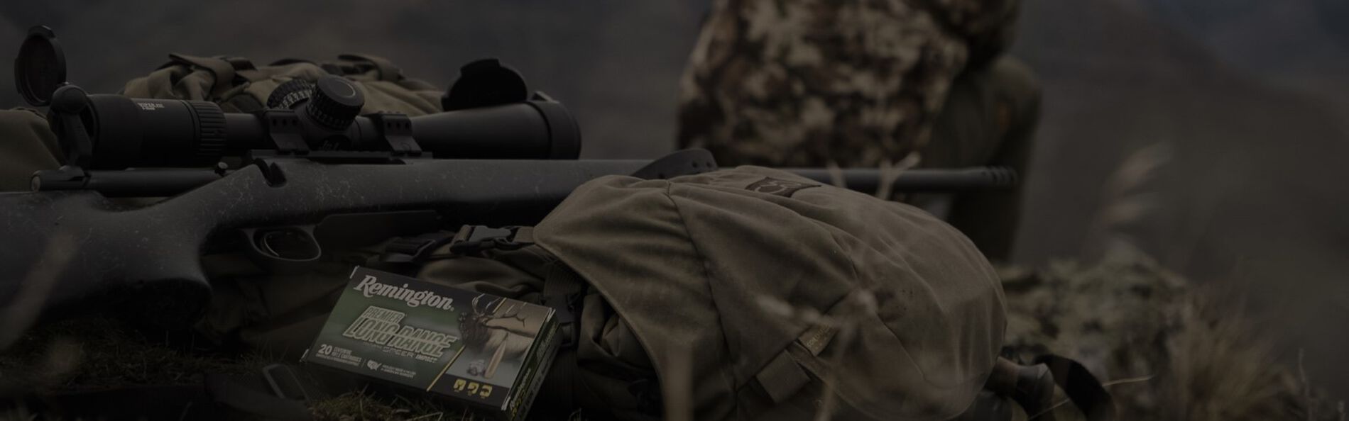 Premier Long Range box laying next to a rifle and backpack with a hunter sitting in the background