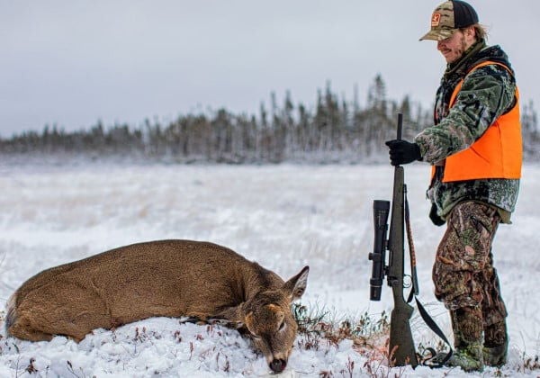 hunter standing in front of a dead deer while holding a rifle